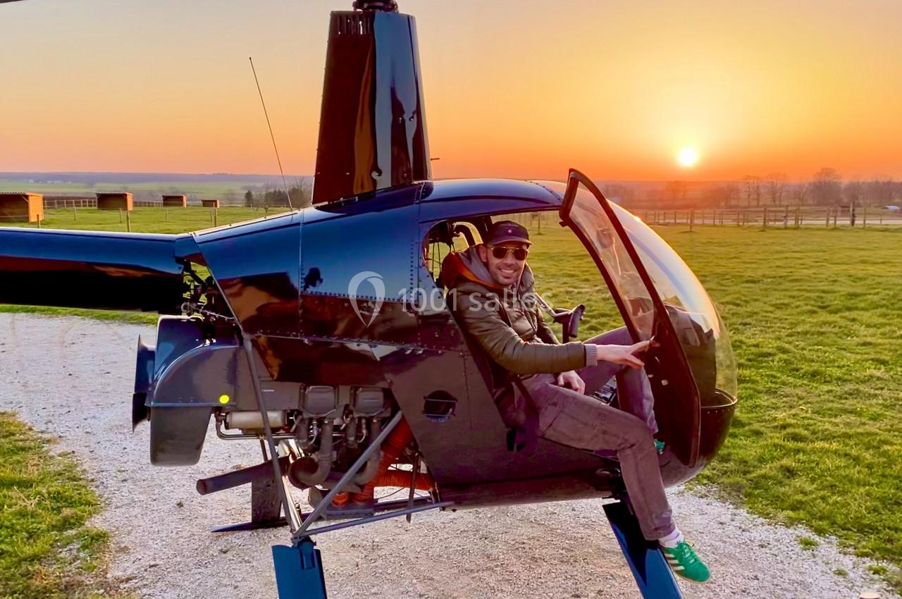 Un homme assis dans un hélicoptère au sol, souriant, avec un coucher de soleil et un champ en arrière-plan.