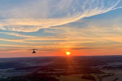 Un homme assis dans un hélicoptère au sol, souriant, avec un coucher de soleil et un champ en arrière-plan.