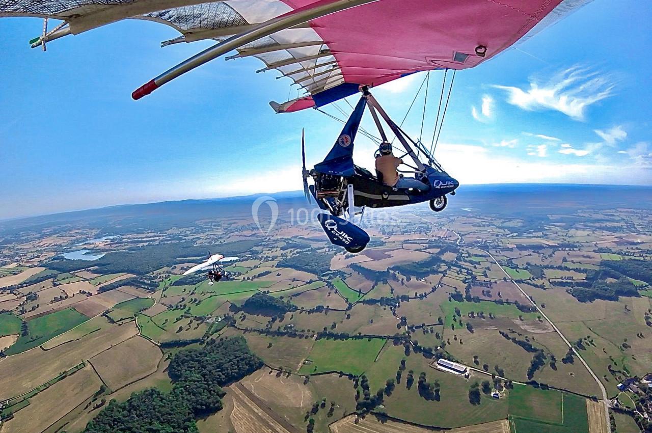 Vue aérienne d'un ULM rose et bleu survolant une campagne verdoyante par temps clair.