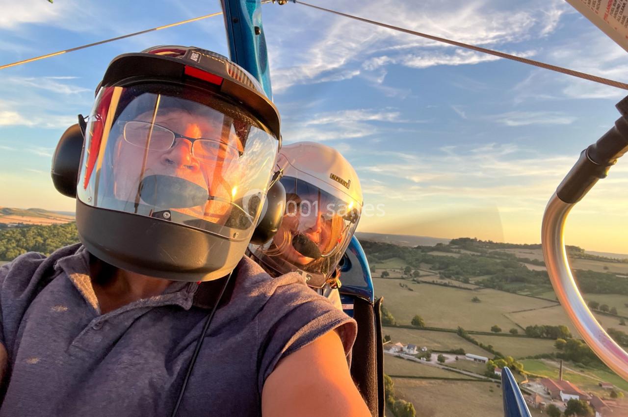 Deux personnes portant des casques volent en ULM au-dessus d'une campagne verdoyante au coucher du soleil.