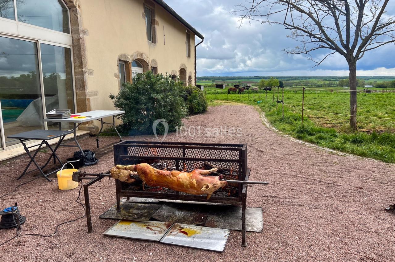 Un cochon entier rôti à la broche devant une maison en pierre, avec un paysage rural en arrière-plan.