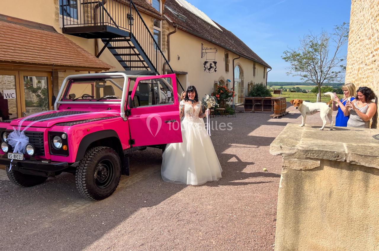 Une mariée en robe blanche sort d'un 4x4 rose décoré, devant un bâtiment rural par temps ensoleillé.