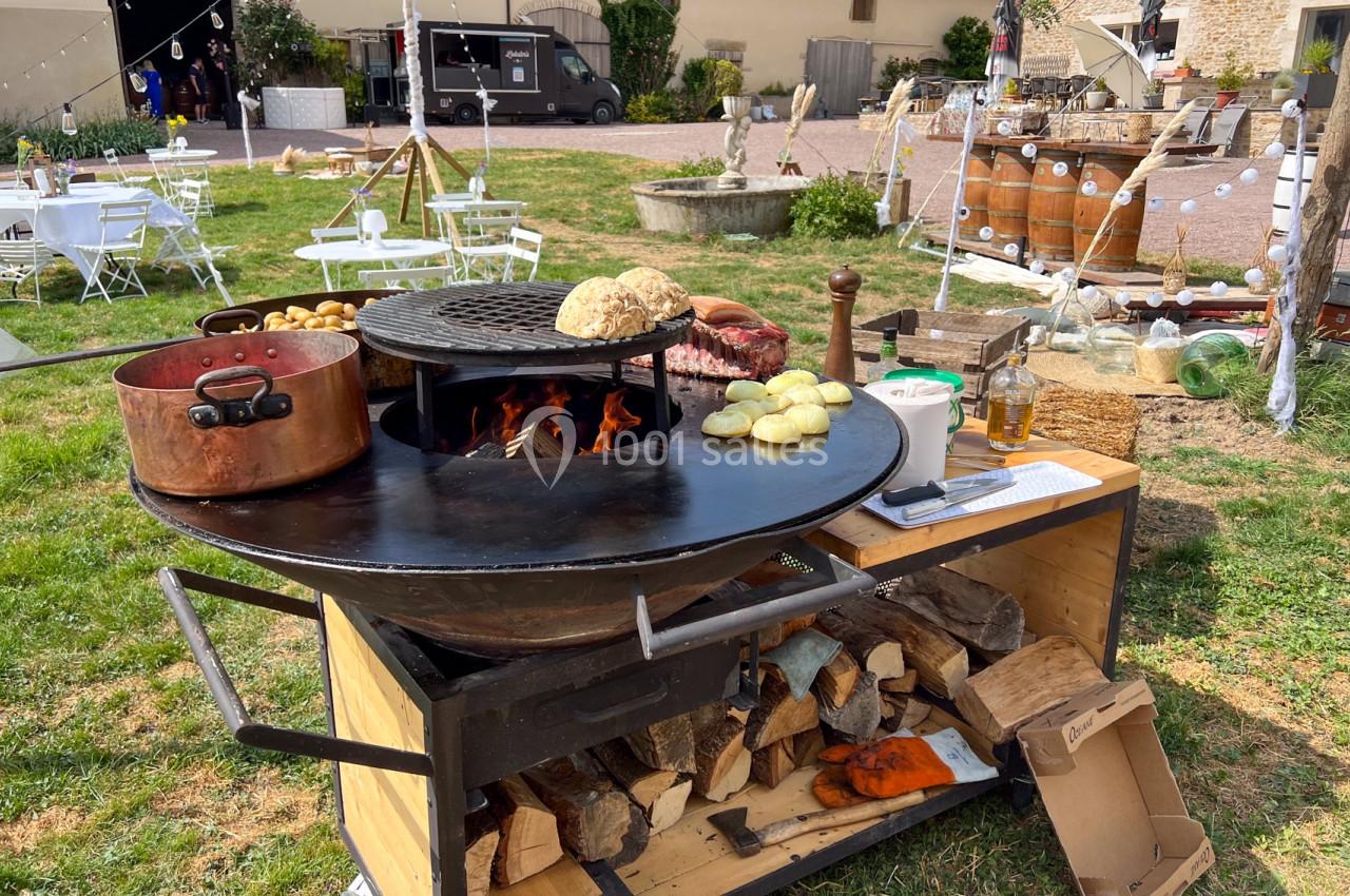 Barbecue circulaire en plein air avec légumes, pain et marmite en cuivre, entouré d'une ambiance festive champêtre.