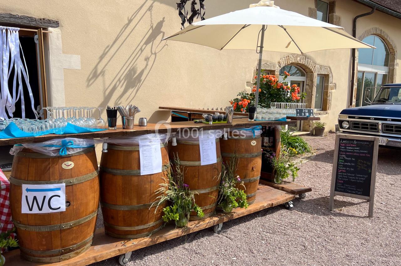 Bar extérieur aménagé avec des tonneaux en bois, décoré de fleurs, sous un parasol près d'un bâtiment en pierre.