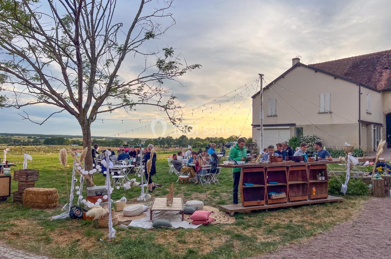 Personnes dînant en extérieur dans une ambiance champêtre, avec guirlandes lumineuses et vue sur la campagne au coucher du…