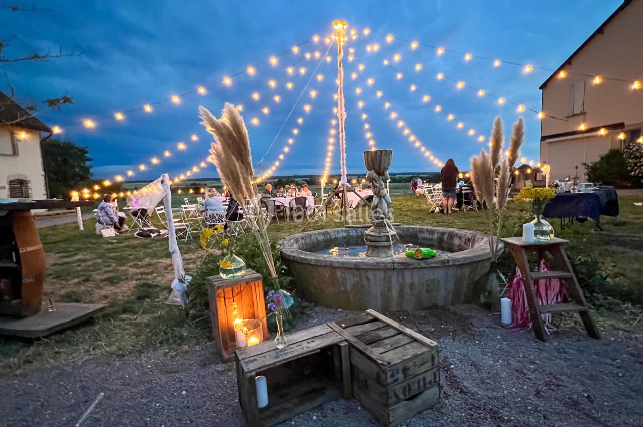 Cour extérieure décorée avec une fontaine centrale, guirlandes lumineuses et tables dressées pour un événement en soirée.