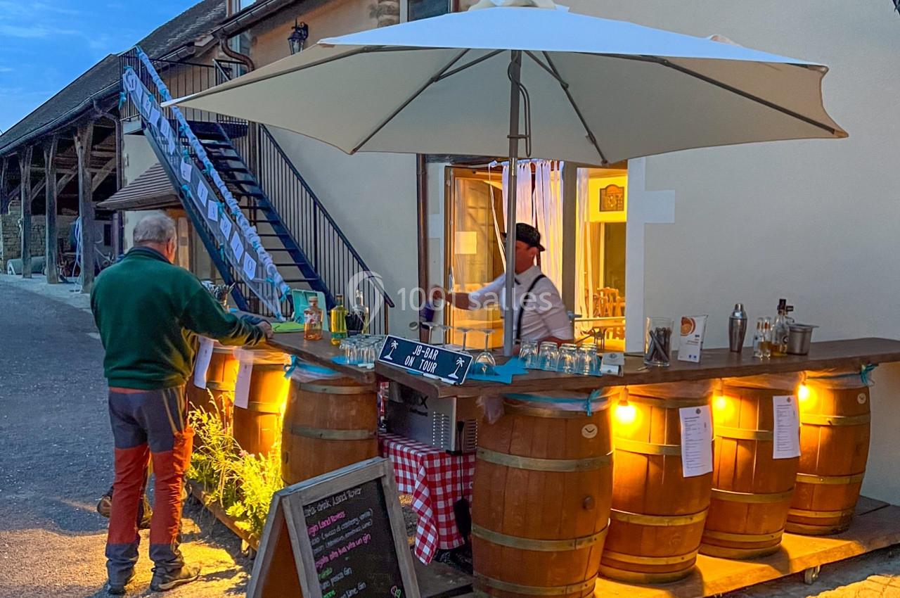 Stand extérieur éclairé en soirée, avec bar en tonneaux, parasol blanc et ardoise de menu devant une maison en pierre.