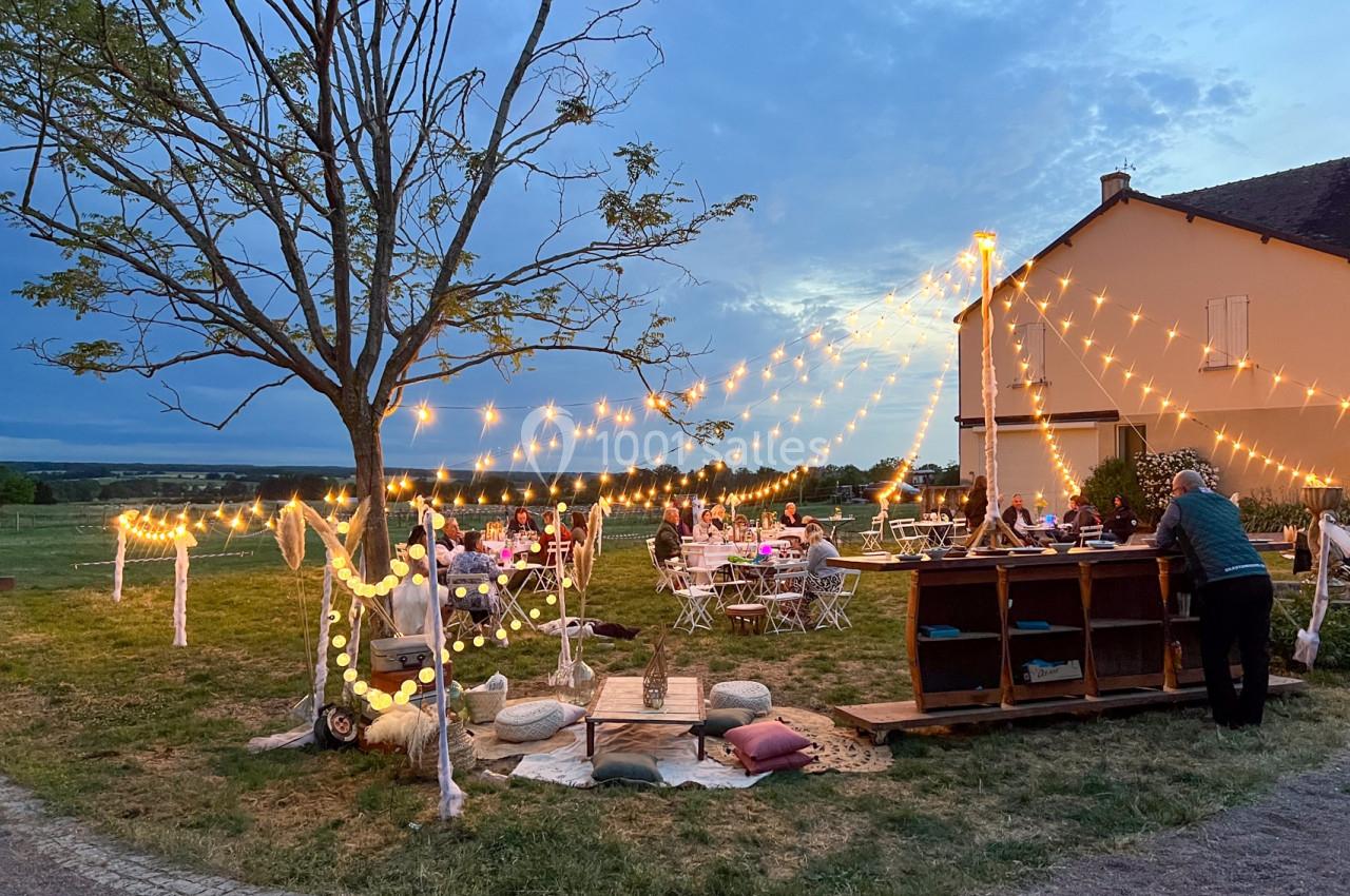 Espace extérieur décoré avec guirlandes lumineuses, tables dressées et bar en bois devant une maison au crépuscule.