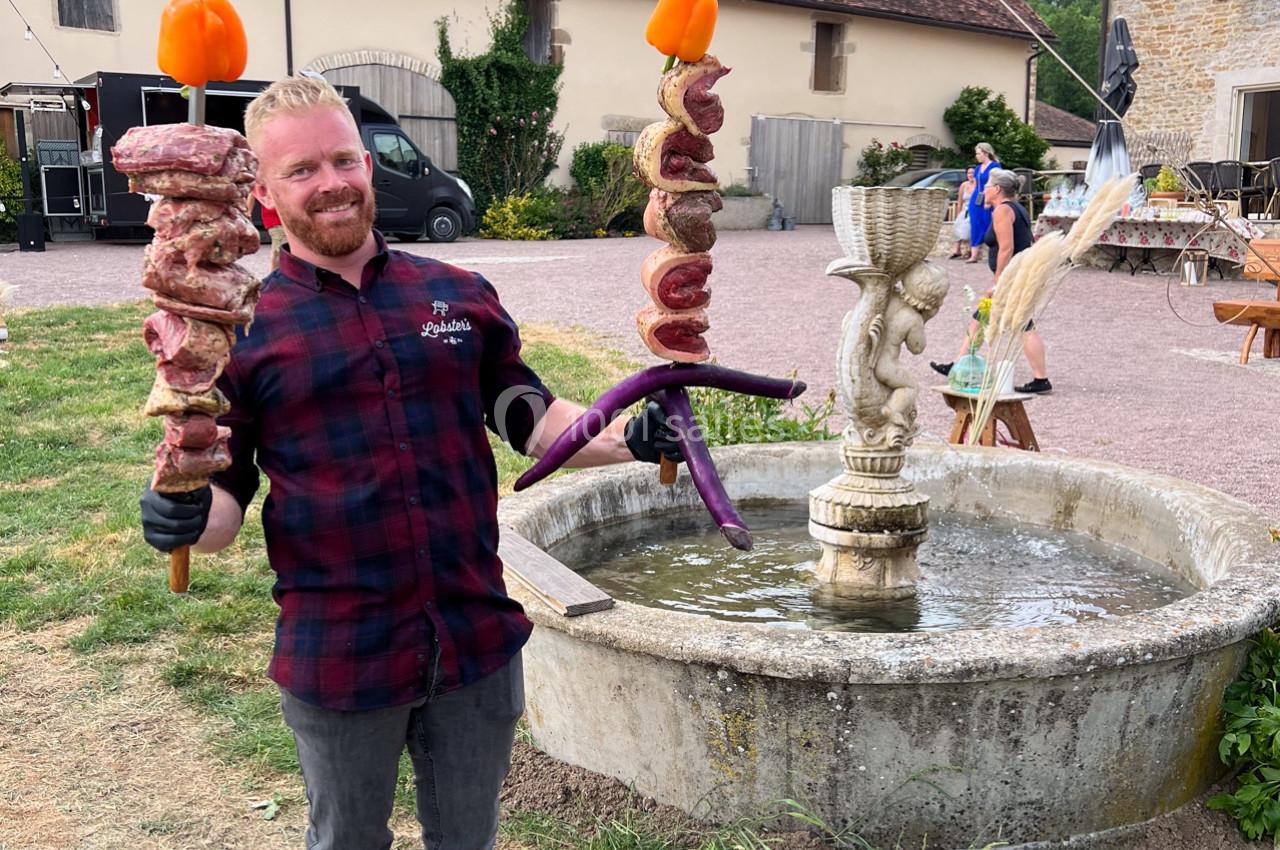 Homme souriant tenant des brochettes géantes devant une fontaine dans un cadre rural.