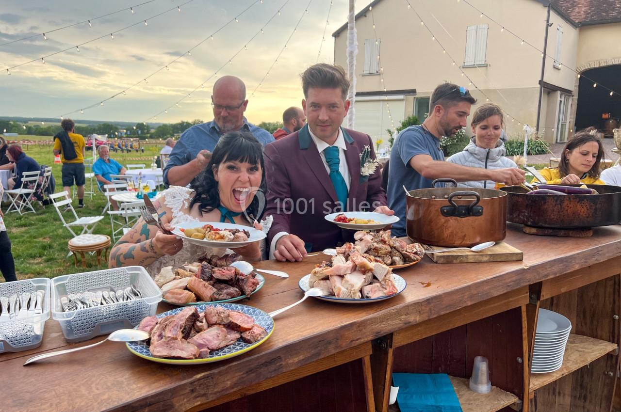 Un couple en tenue de mariage se sert à un buffet en plein air, entouré de convives et de plats variés.