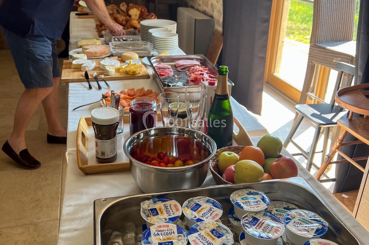 Un buffet de petit-déjeuner avec yaourts, fruits, viennoiseries, charcuterie et boissons, dans une salle lumineuse.