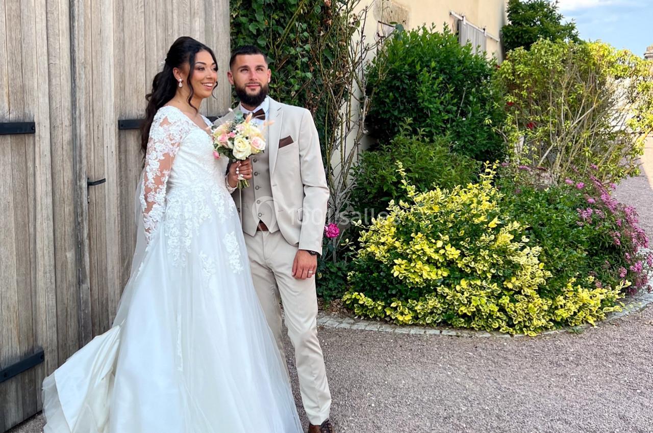 Un couple en tenue de mariage pose devant un bâtiment en pierre entouré de végétation.