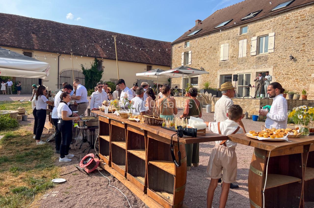 Groupe de personnes rassemblées dans une cour ensoleillée devant un bâtiment en pierre, autour de tables de buffet.