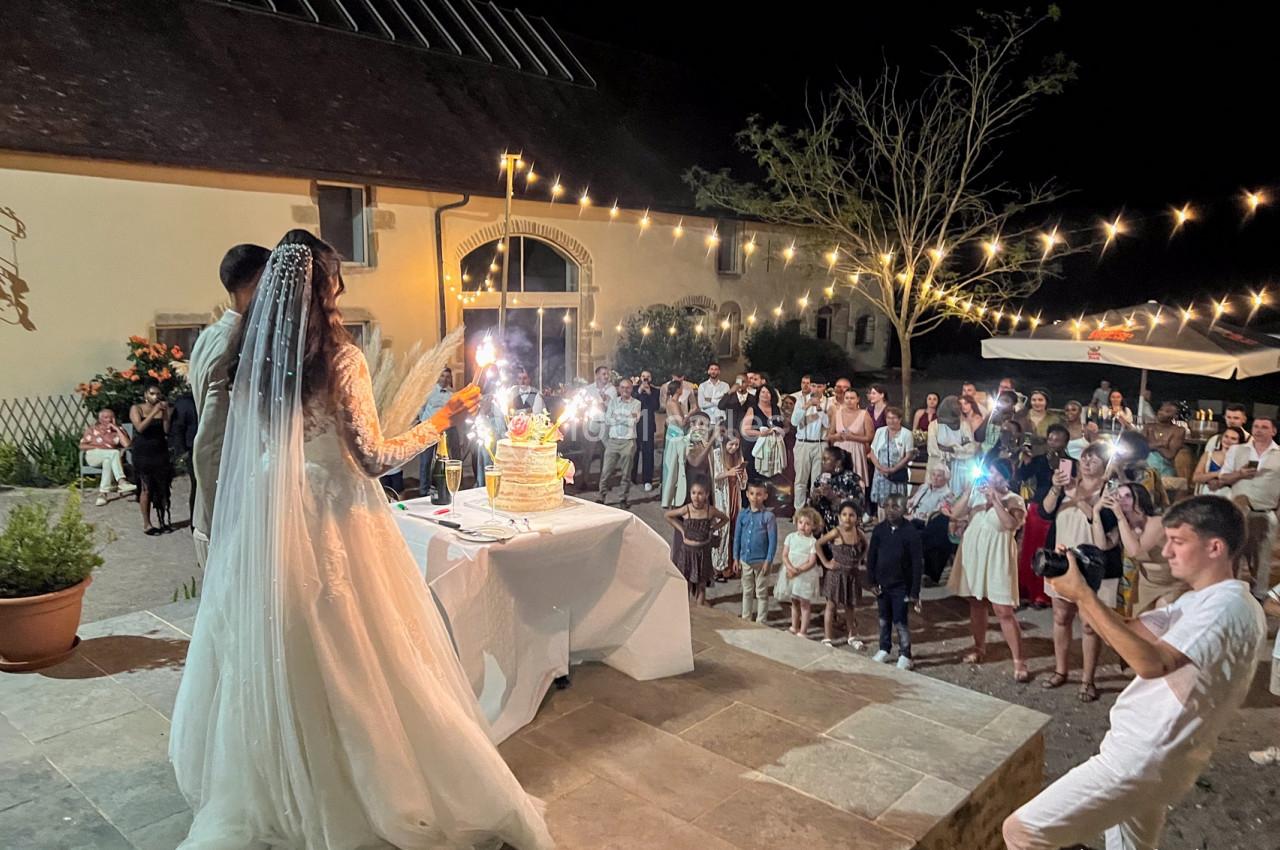 Un couple de mariés coupe un gâteau de mariage illuminé, entouré d'invités en soirée sous des guirlandes lumineuses.