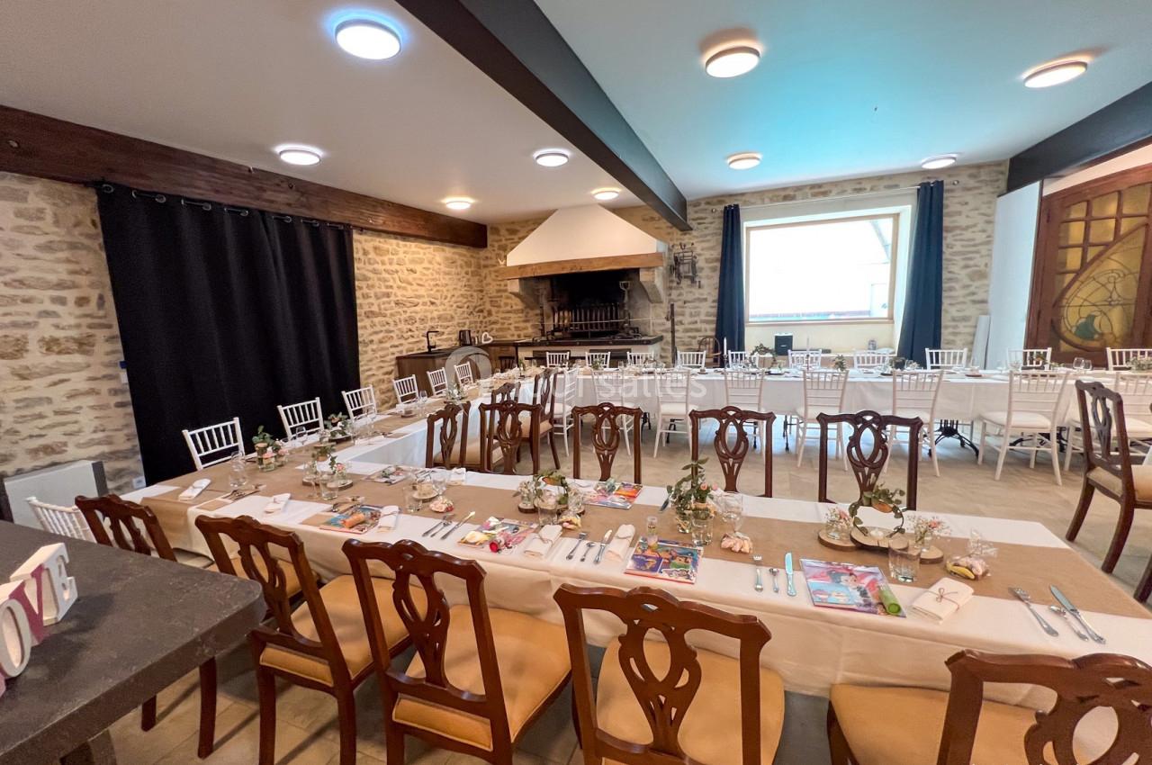 Salle de réception avec tables dressées, chaises en bois et murs en pierre, éclairée par des plafonniers et une grande…