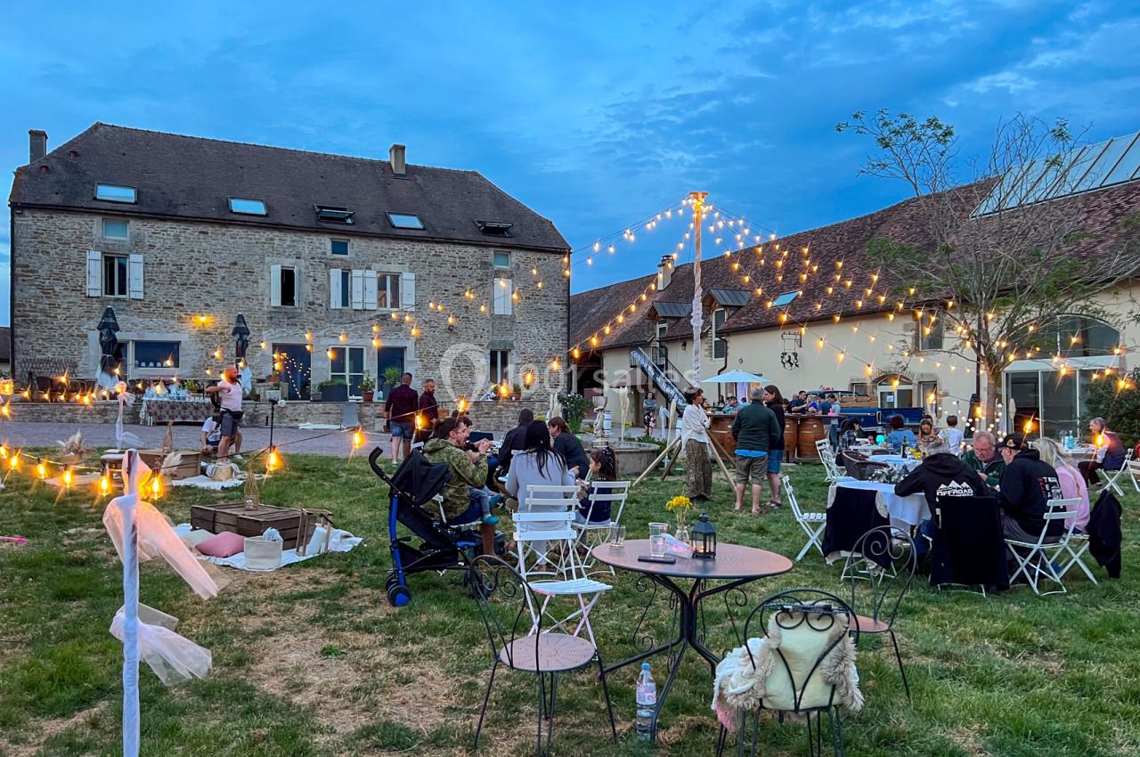 Personnes rassemblées dans un jardin en soirée, entourées de guirlandes lumineuses et de bâtiments en pierre.