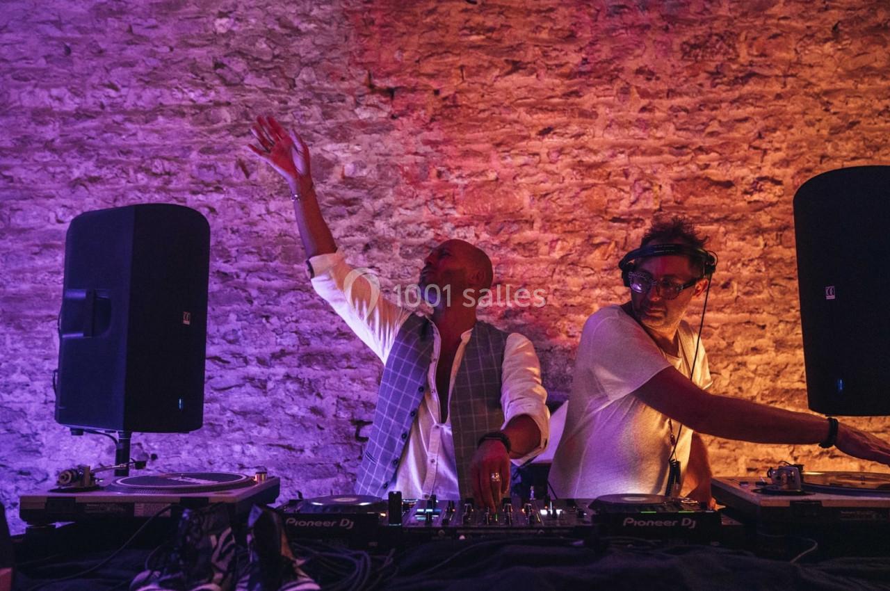 Deux DJ mixent de la musique sur une scène éclairée, entourés de haut-parleurs et d'un mur en pierre.