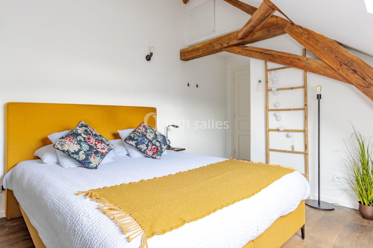 Chambre lumineuse avec lit double, tête de lit jaune, poutres apparentes, échelle décorative et plante verte.