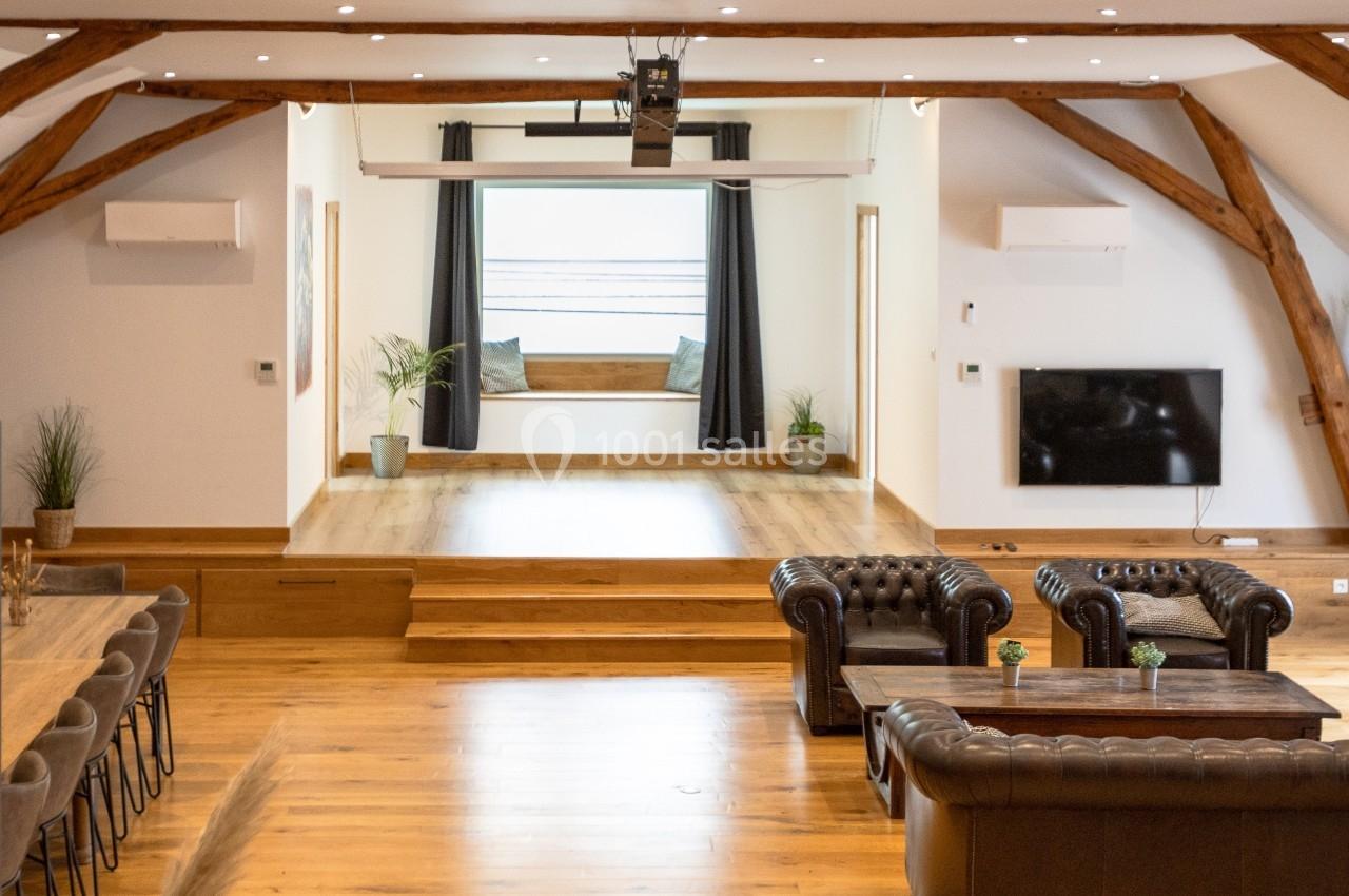 Salle lumineuse avec parquet, canapés en cuir, table en bois, et espace surélevé avec fenêtre et rideaux noirs.