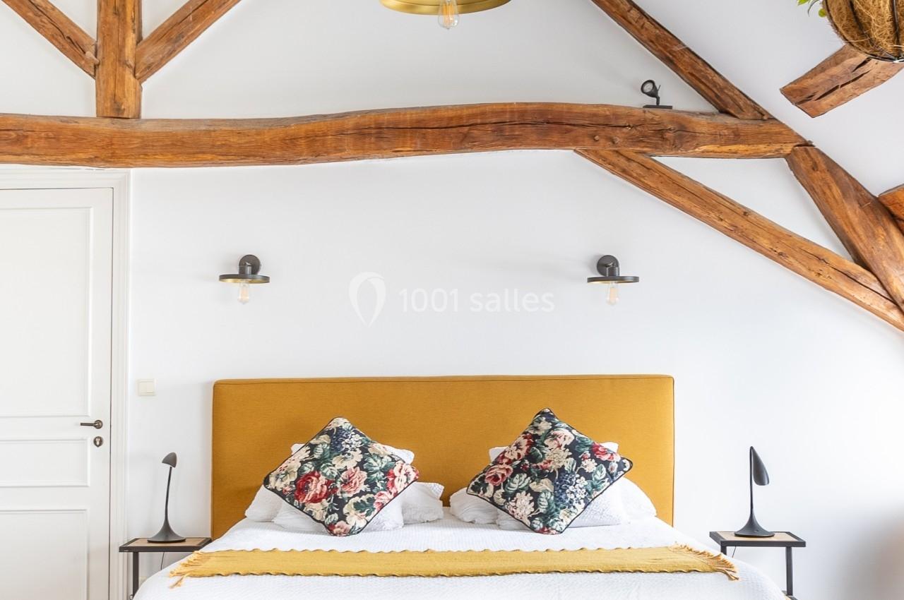 Lit double avec tête de lit jaune moutarde, poutres apparentes en bois, suspension et décoration murale dans une chambre…