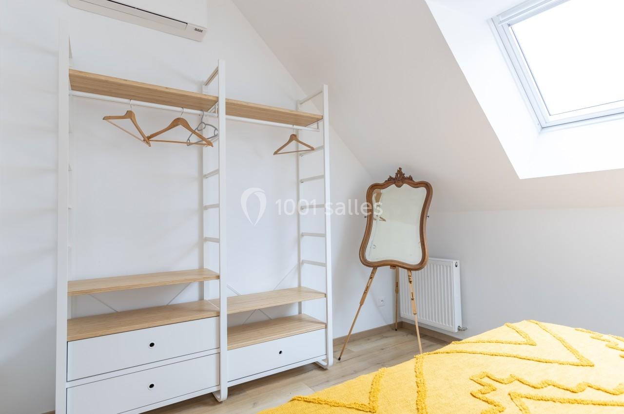 Armoire ouverte en bois et métal avec cintres, miroir ancien sur pied et lumière naturelle d'une fenêtre de toit.