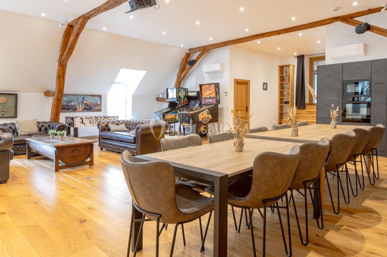 Salle lumineuse avec table en bois, chaises en cuir, canapés Chesterfield, flippers et poutres apparentes.