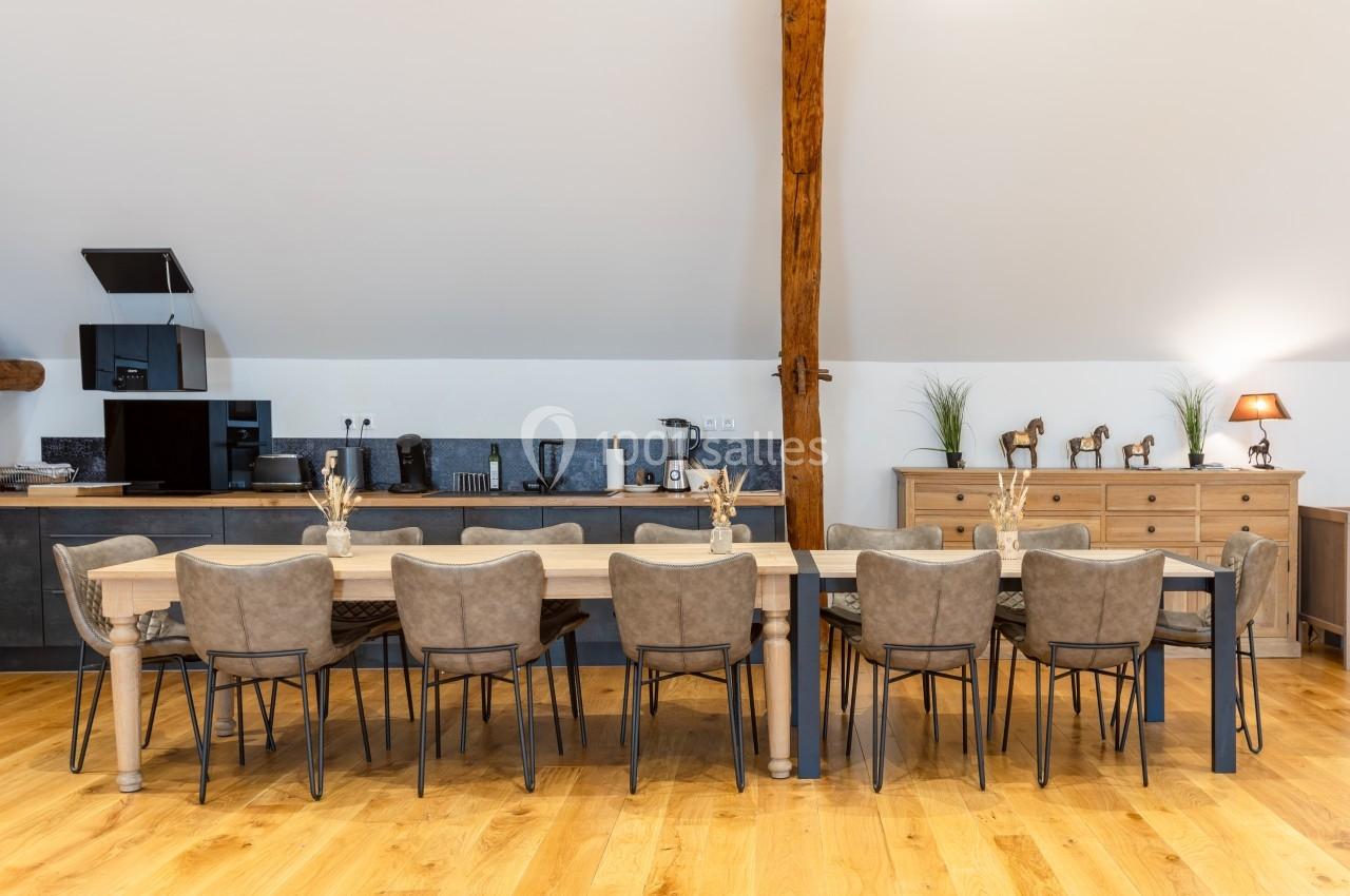 Grande table en bois entourée de chaises grises dans une salle à manger lumineuse avec cuisine ouverte et décoration sobre.