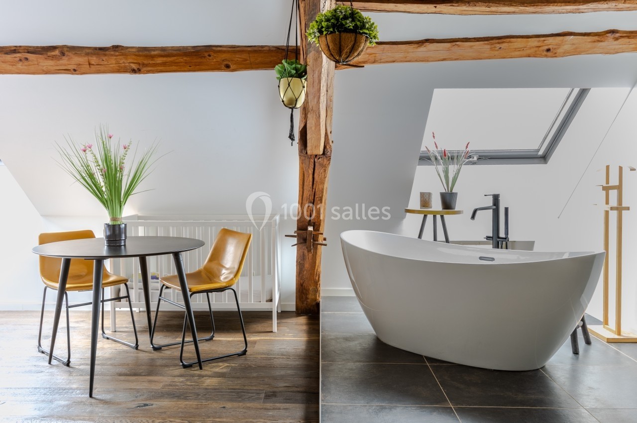 Location salle Souhey (Côte-d'Or) - La Maison de Souhey #48 Salle de bain lumineuse avec baignoire îlot, table ronde, chaises en cuir et poutres apparentes sous un toit mansardé.