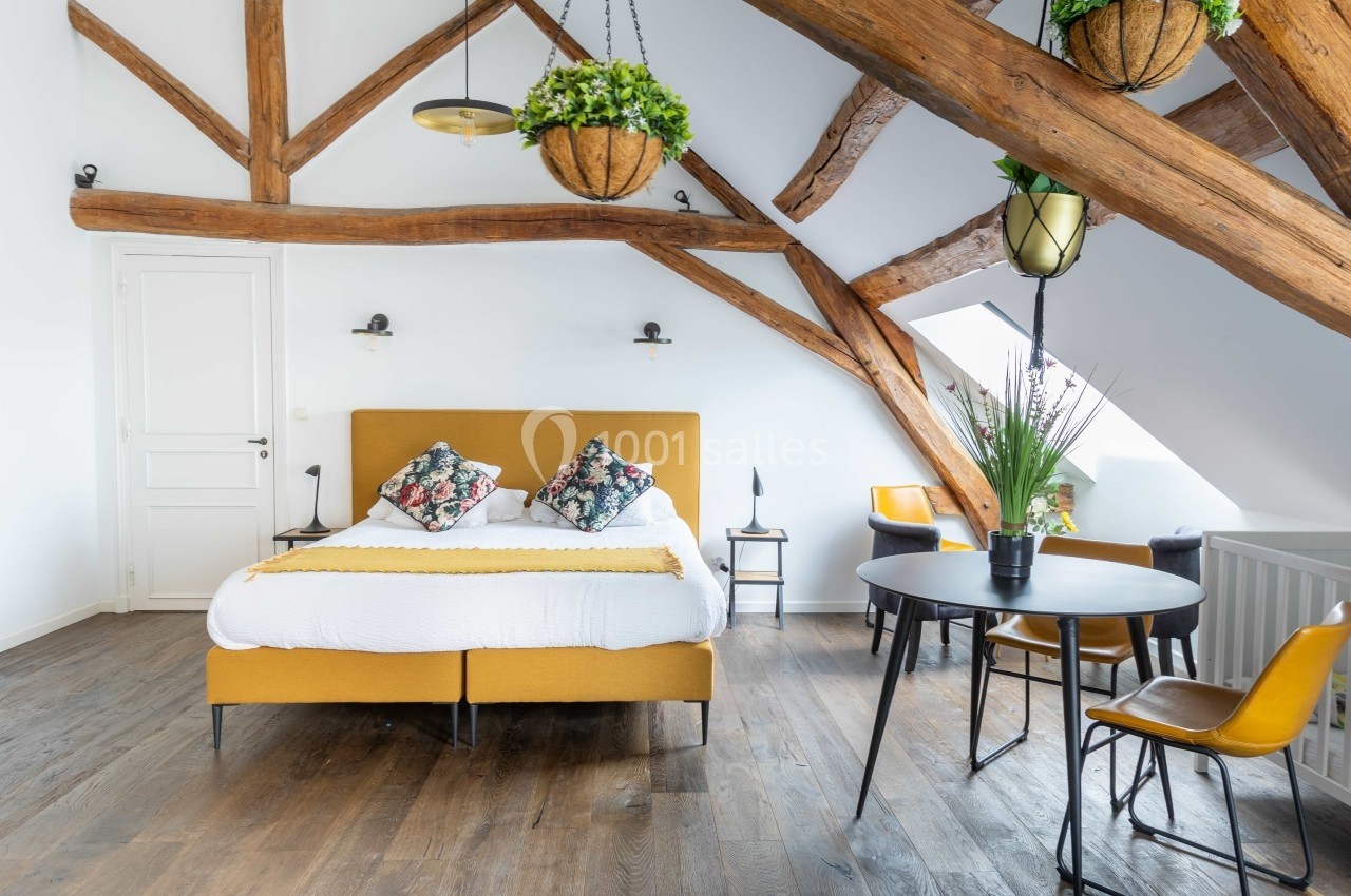 Location salle Souhey (Côte-d'Or) - La Maison de Souhey #42 Chambre mansardée lumineuse avec poutres apparentes, lit double, table ronde, chaises et plantes suspendues.