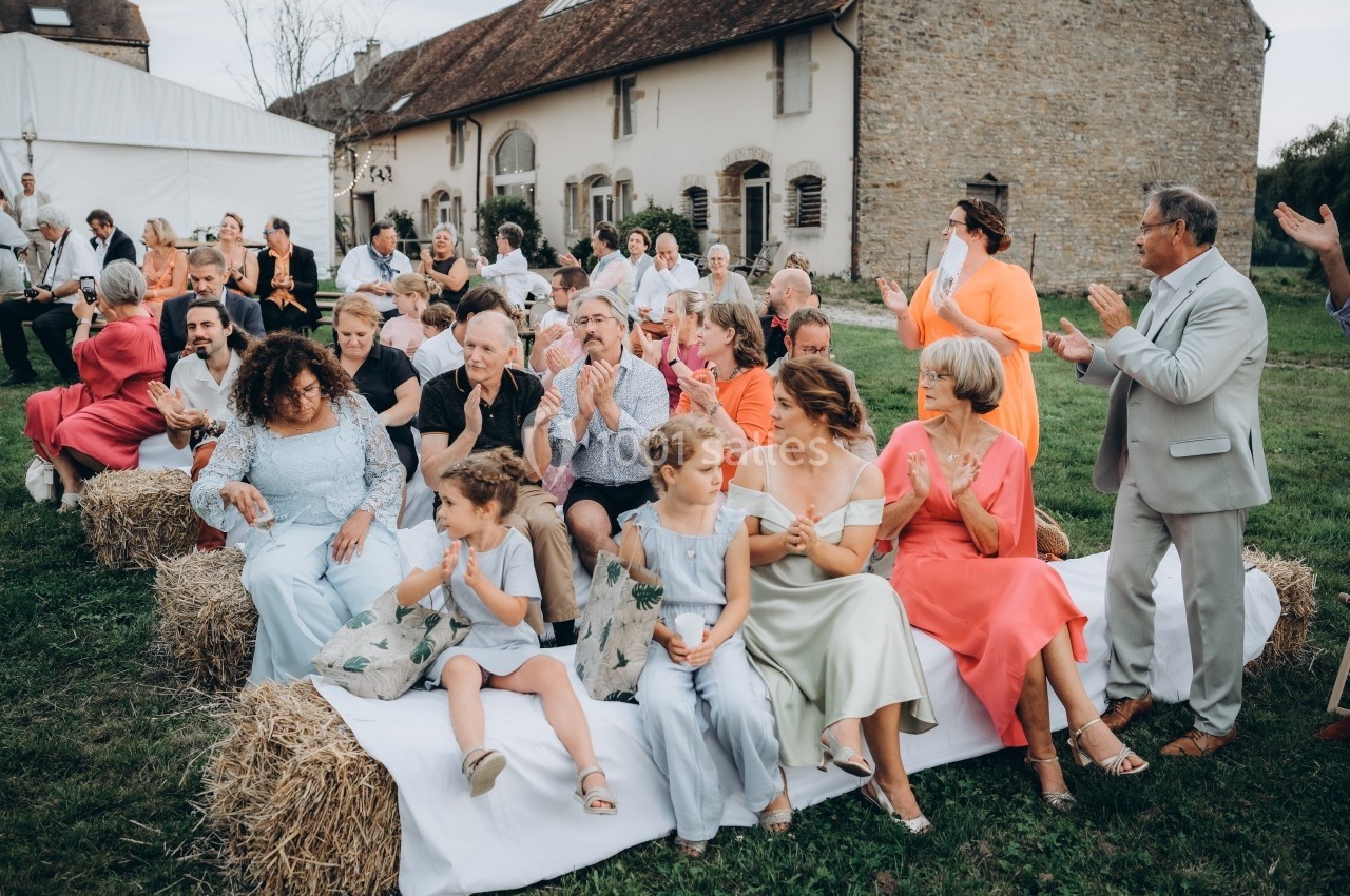 Location salle Souhey (Côte-d'Or) - La Maison de Souhey #76 Des invités assis sur des bottes de foin applaudissent lors d'un événement en plein air près d'un bâtiment en pierre.