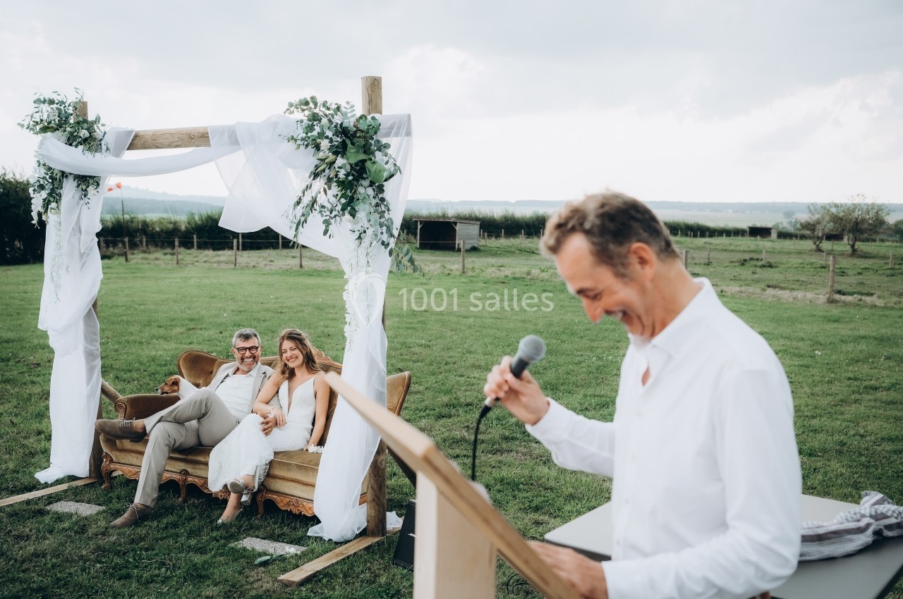 Location salle Souhey (Côte-d'Or) - La Maison de Souhey #78 Un couple assis sous une arche décorée dans un champ, écoutant un homme parlant au micro depuis un pupitre.