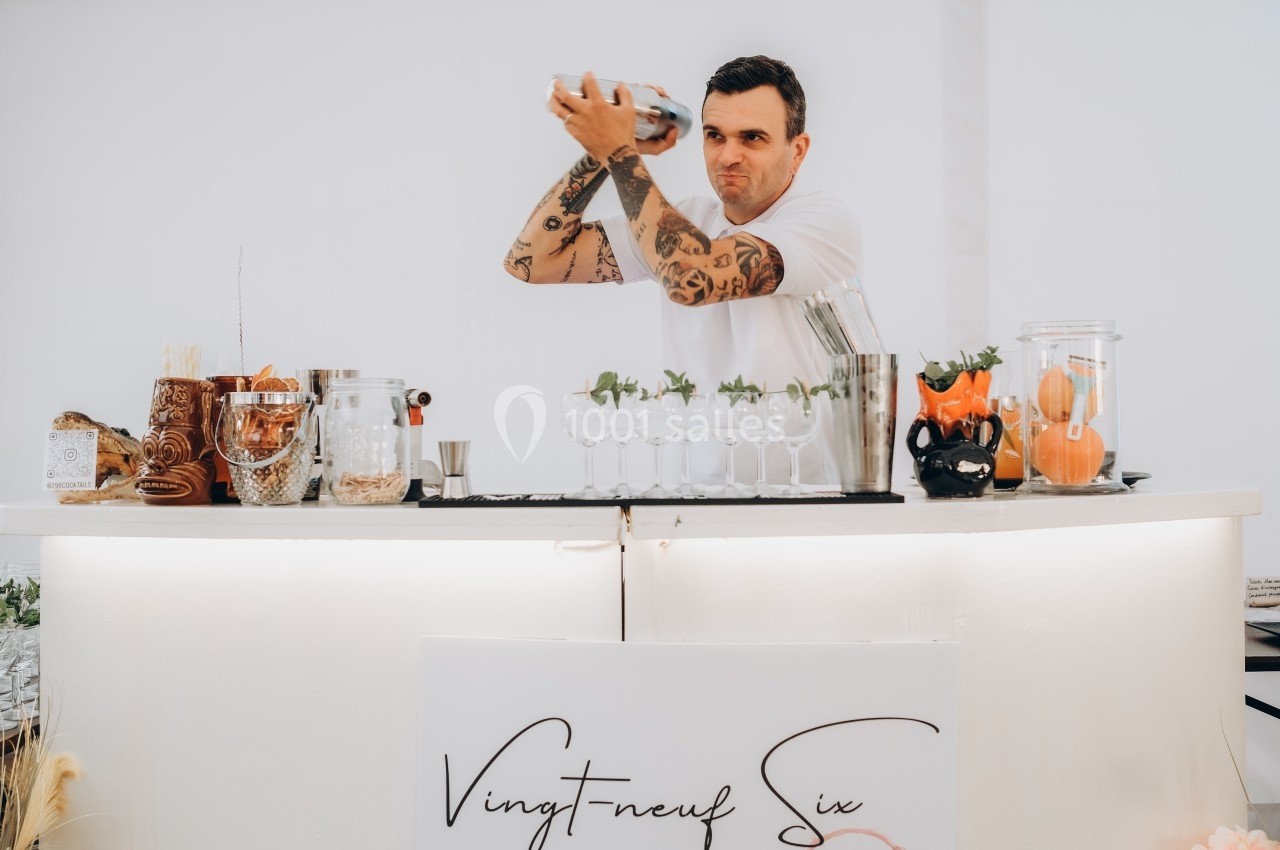 Location salle Souhey (Côte-d'Or) - La Maison de Souhey #80 Un barman tatoué prépare un cocktail en secouant un shaker derrière un comptoir blanc décoré de verres et d'ingrédients.