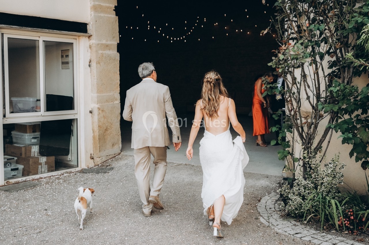 Location salle Souhey (Côte-d'Or) - La Maison de Souhey #82 Une mariée en robe blanche marche avec un homme en costume beige et un petit chien vers une entrée décorée de guirlandes…