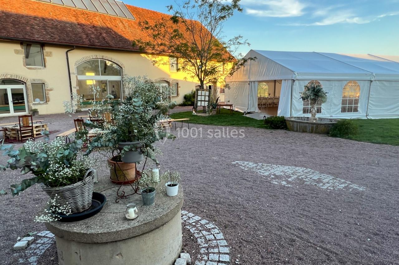 Cour d'une maison de campagne avec un puits fleuri, une tente blanche et une bâtisse en pierre sous un ciel dégagé.