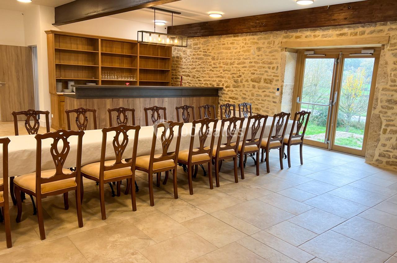 Salle avec une longue table dressée, entourée de chaises en bois, murs en pierre et grande porte vitrée donnant sur l…
