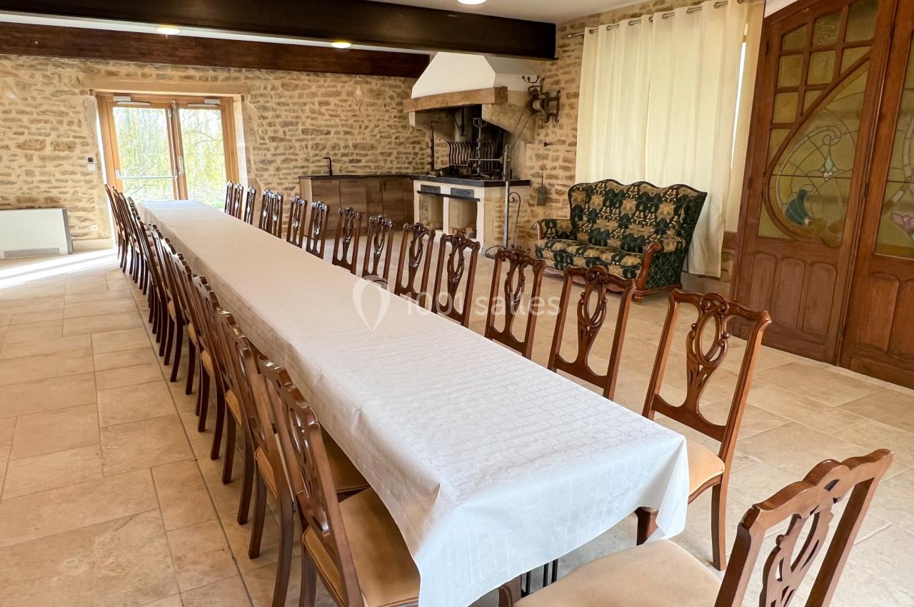 Grande salle avec une longue table recouverte d'une nappe blanche, entourée de chaises en bois, et une cheminée en pierre.