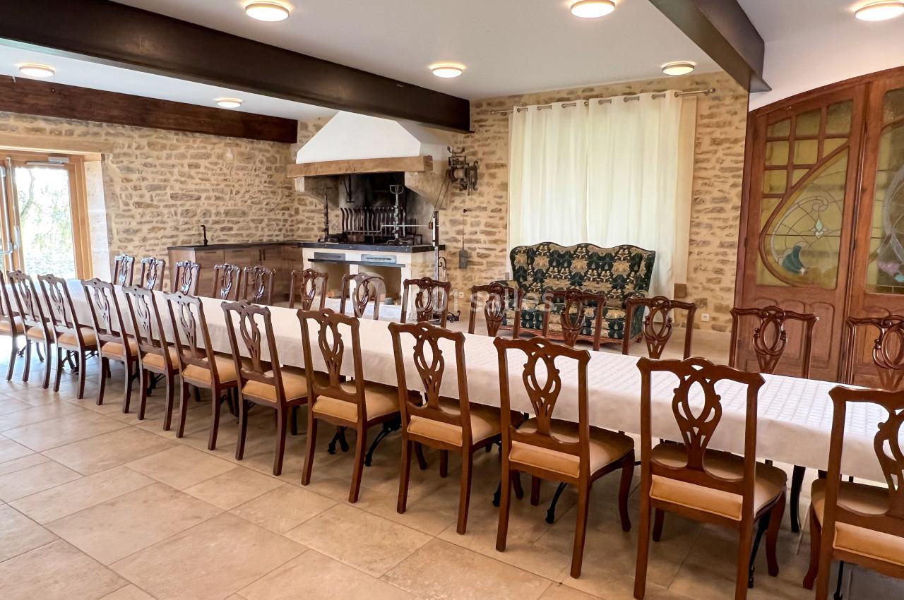 Grande salle avec une longue table dressée, entourée de chaises en bois, et un mur en pierre avec une cheminée.