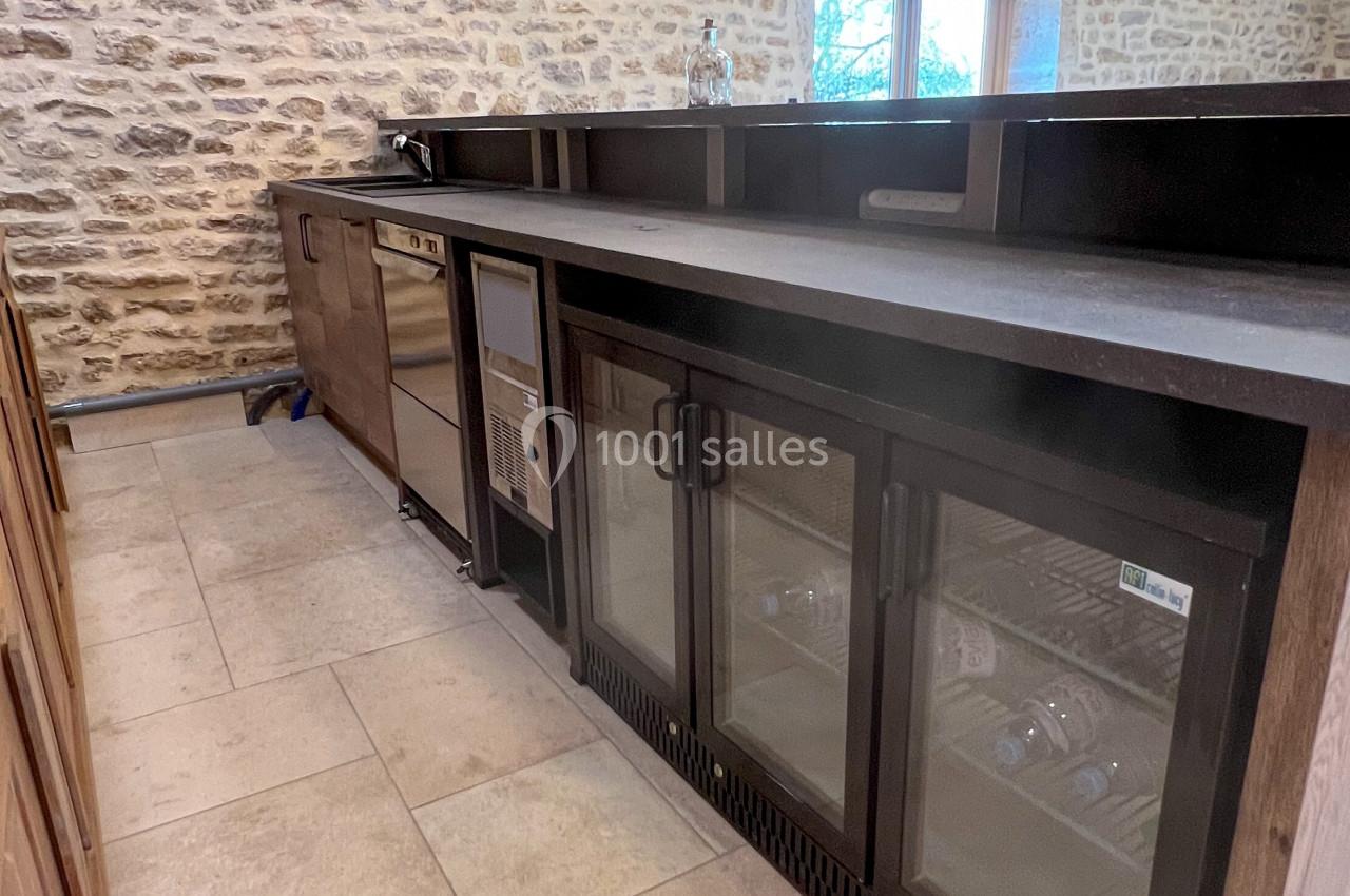 Cuisine avec comptoir en bois foncé, murs en pierre apparente, sol carrelé beige et réfrigérateurs vitrés intégrés.