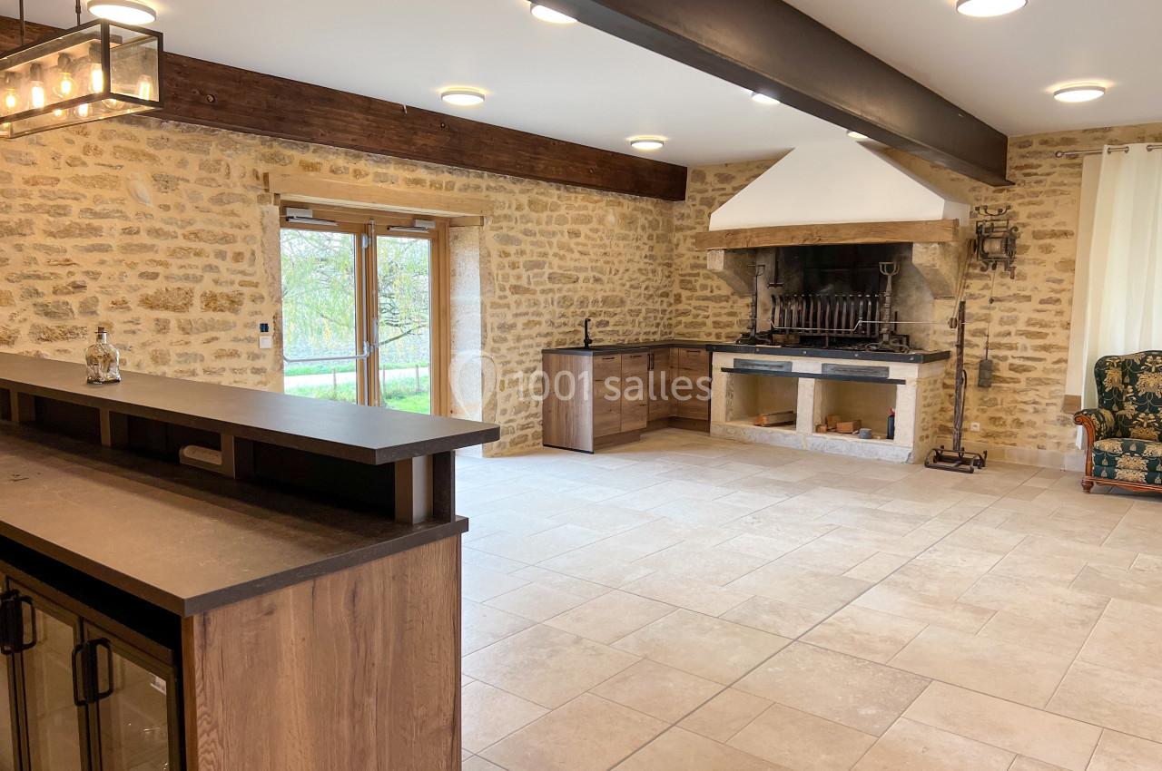 Cuisine ouverte avec murs en pierre, poutres apparentes, cheminée rustique et mobilier en bois moderne.