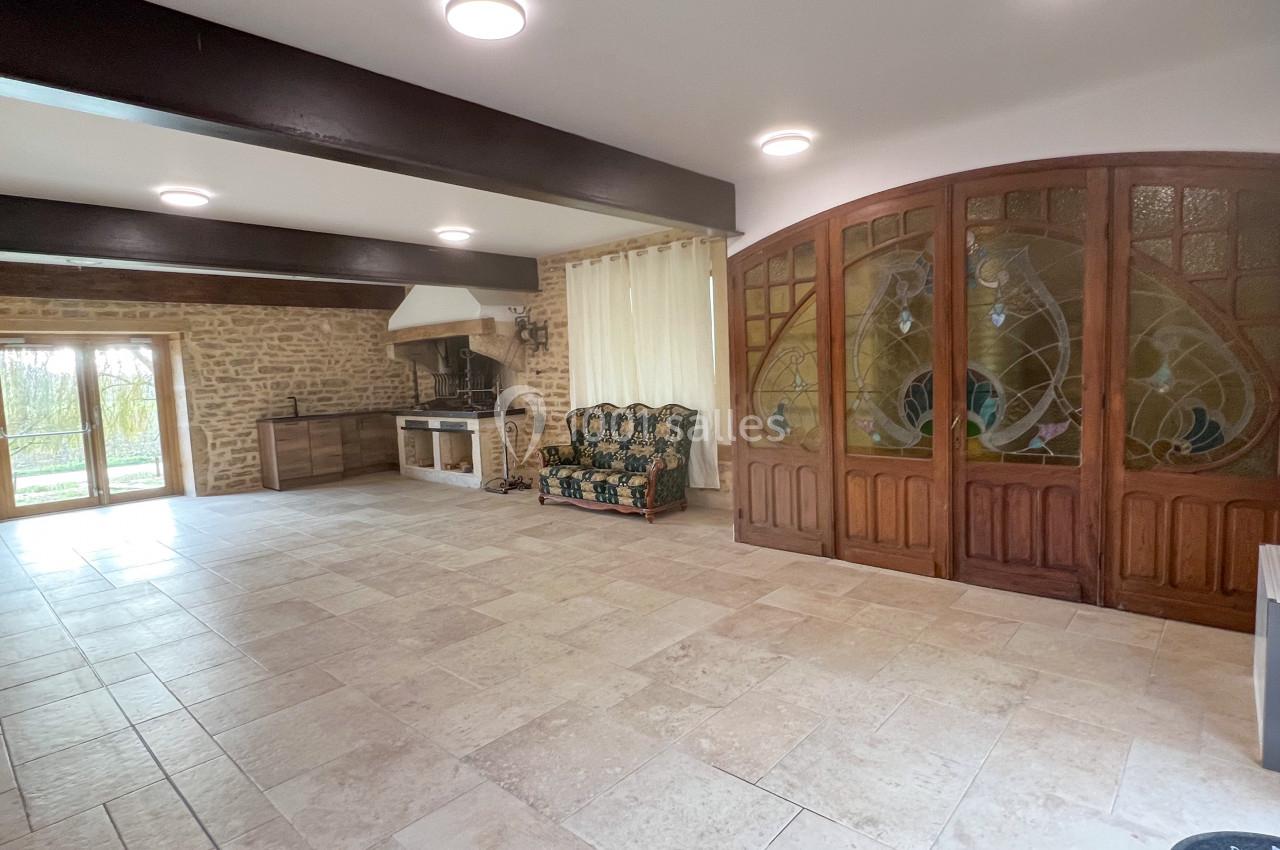 Salle spacieuse avec murs en pierre, poutres apparentes, portes vitrées en bois et cheminée intégrée.