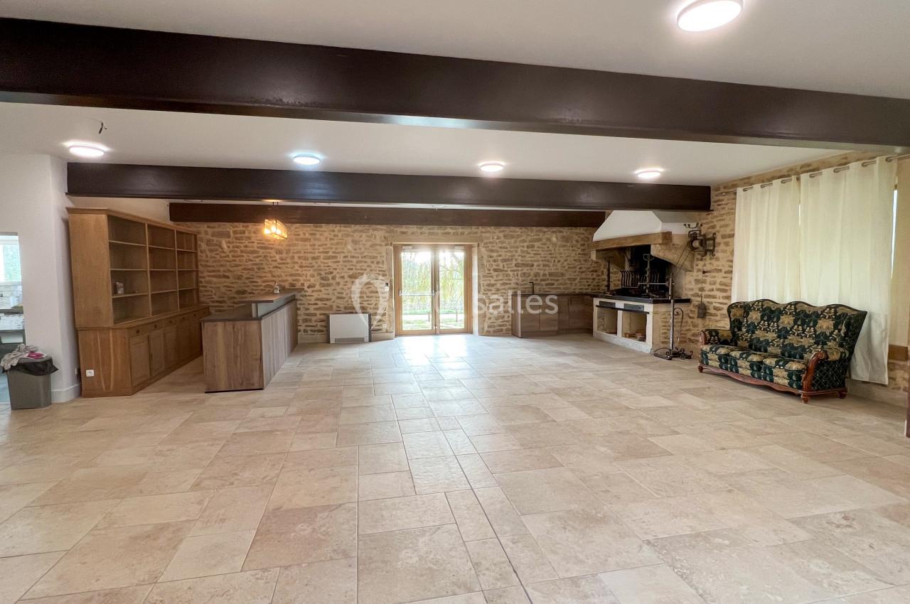 Salle spacieuse avec murs en pierre, carrelage clair, cheminée, canapé et grandes portes vitrées donnant sur l'extérieur.