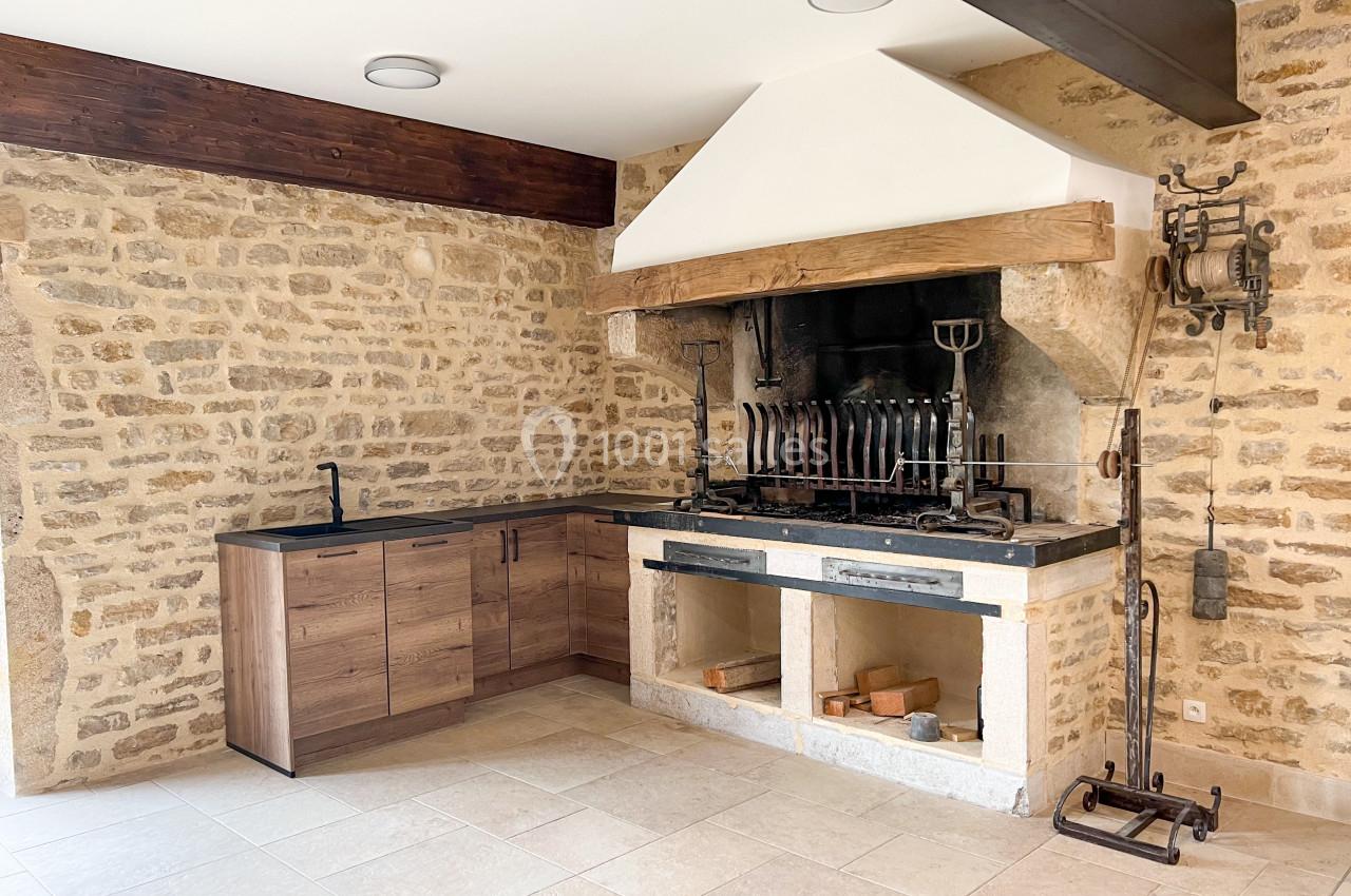 Cuisine d'été avec murs en pierre, cheminée équipée d'une rôtissoire et meubles en bois avec évier intégré.