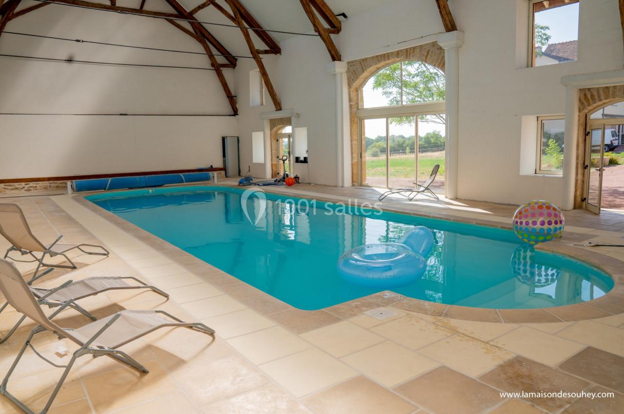 Piscine intérieure lumineuse avec chaises longues, bouées et grandes baies vitrées donnant sur un paysage verdoyant.