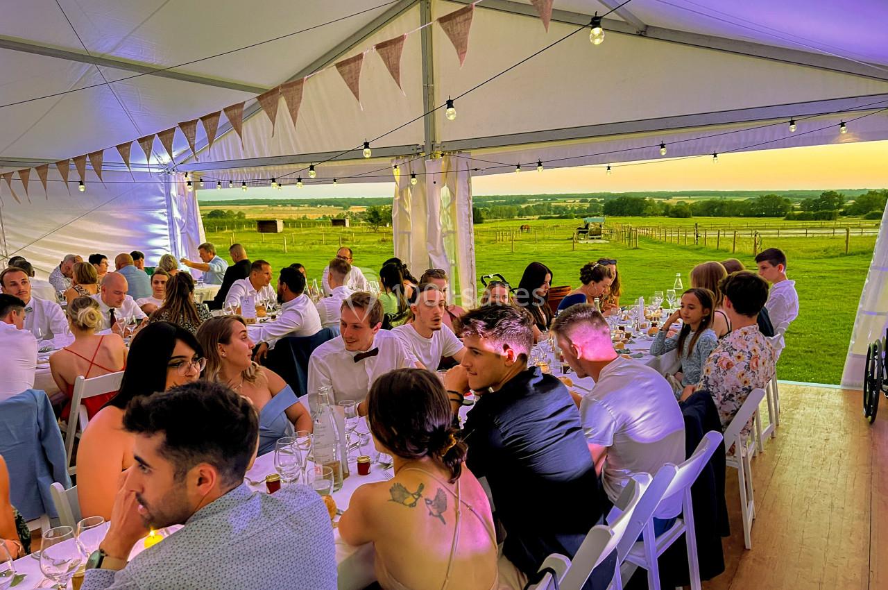 Des invités assis à des tables dans une tente décorée, profitant d'un repas avec vue sur un paysage champêtre au coucher du…