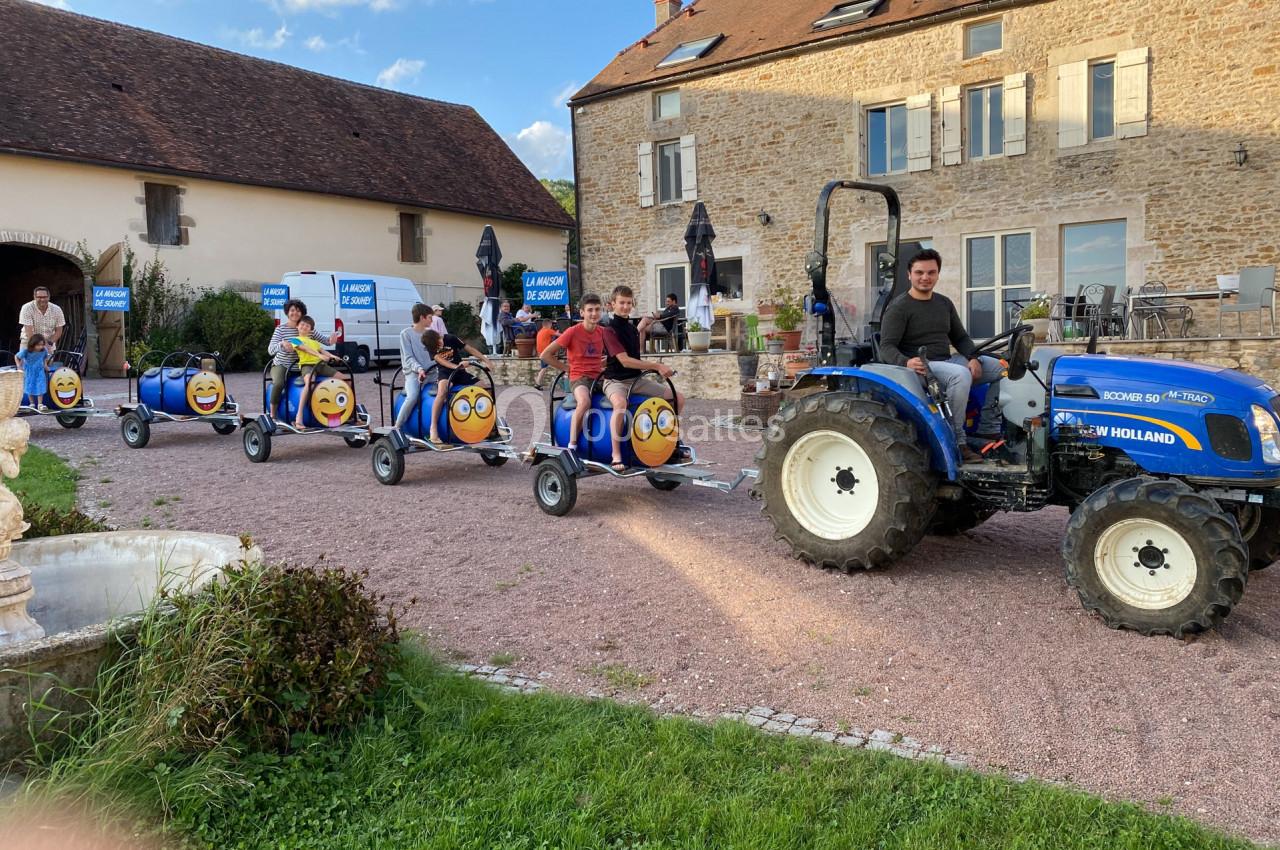 Un tracteur bleu tire un petit train de barils décorés avec des visages souriants, devant des bâtiments en pierre.