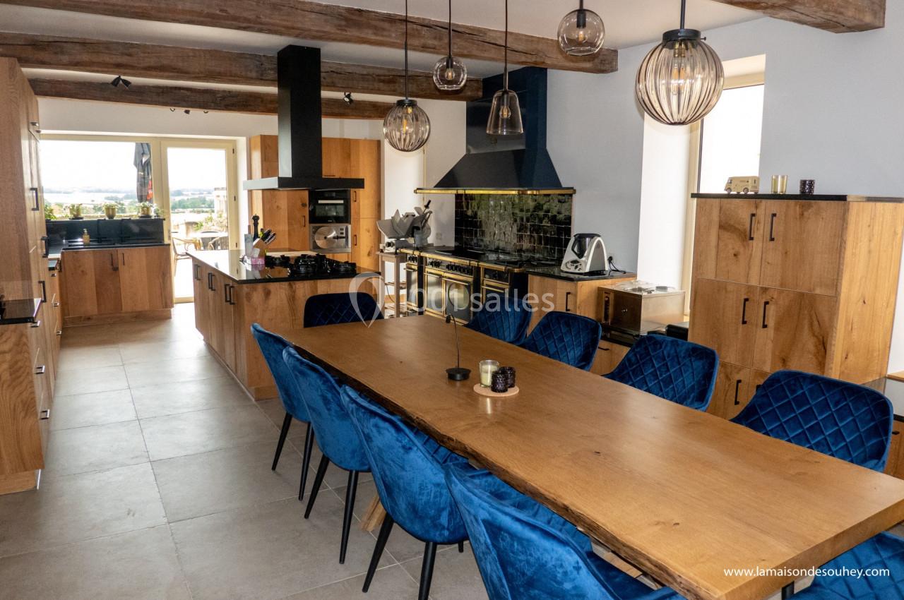 Cuisine spacieuse avec table en bois, chaises bleues, meubles en bois, poutres apparentes et vue sur l'extérieur.