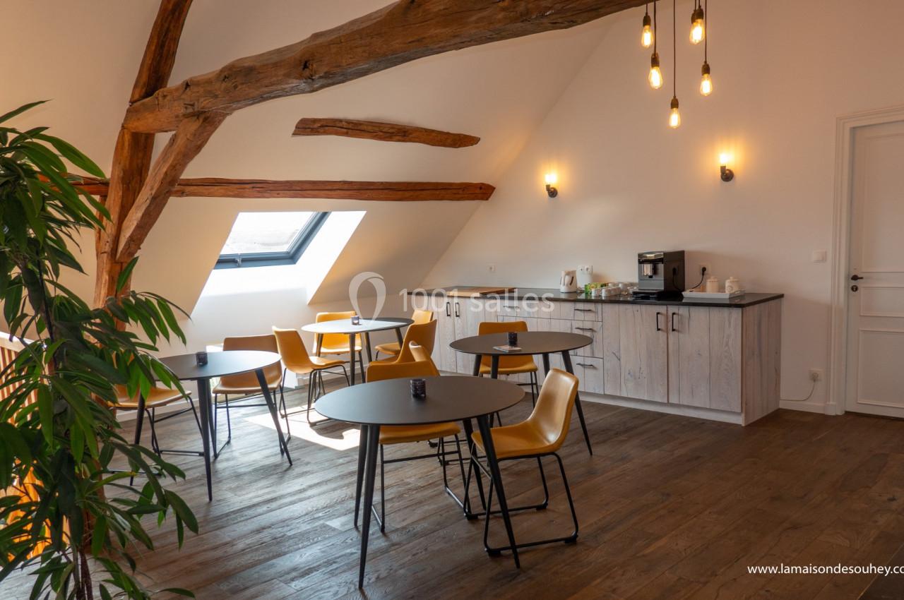 Salle lumineuse sous combles avec tables, chaises jaunes, poutres apparentes, kitchenette et plantes décoratives.