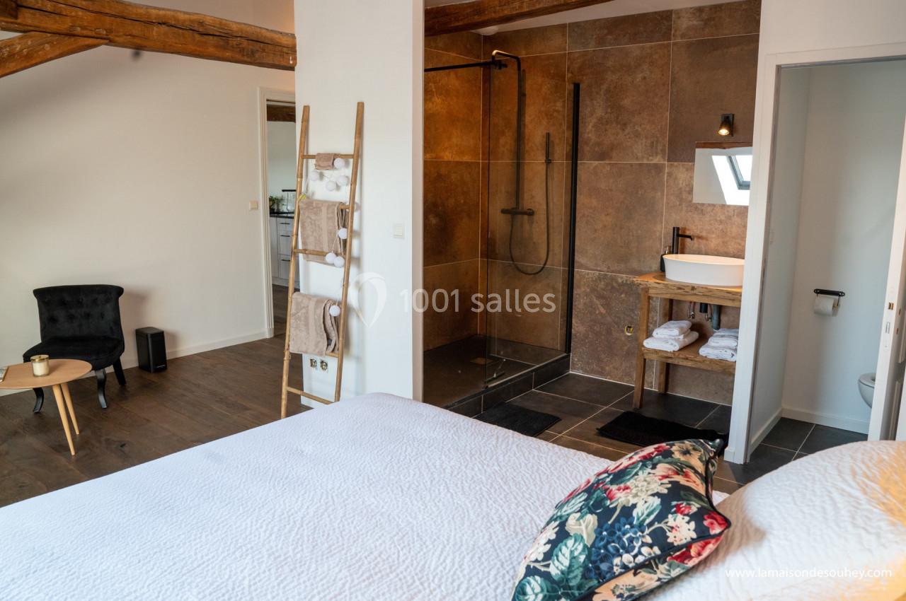 Chambre lumineuse avec lit double, coin salon, salle de bain ouverte avec douche à l'italienne et lavabo moderne.