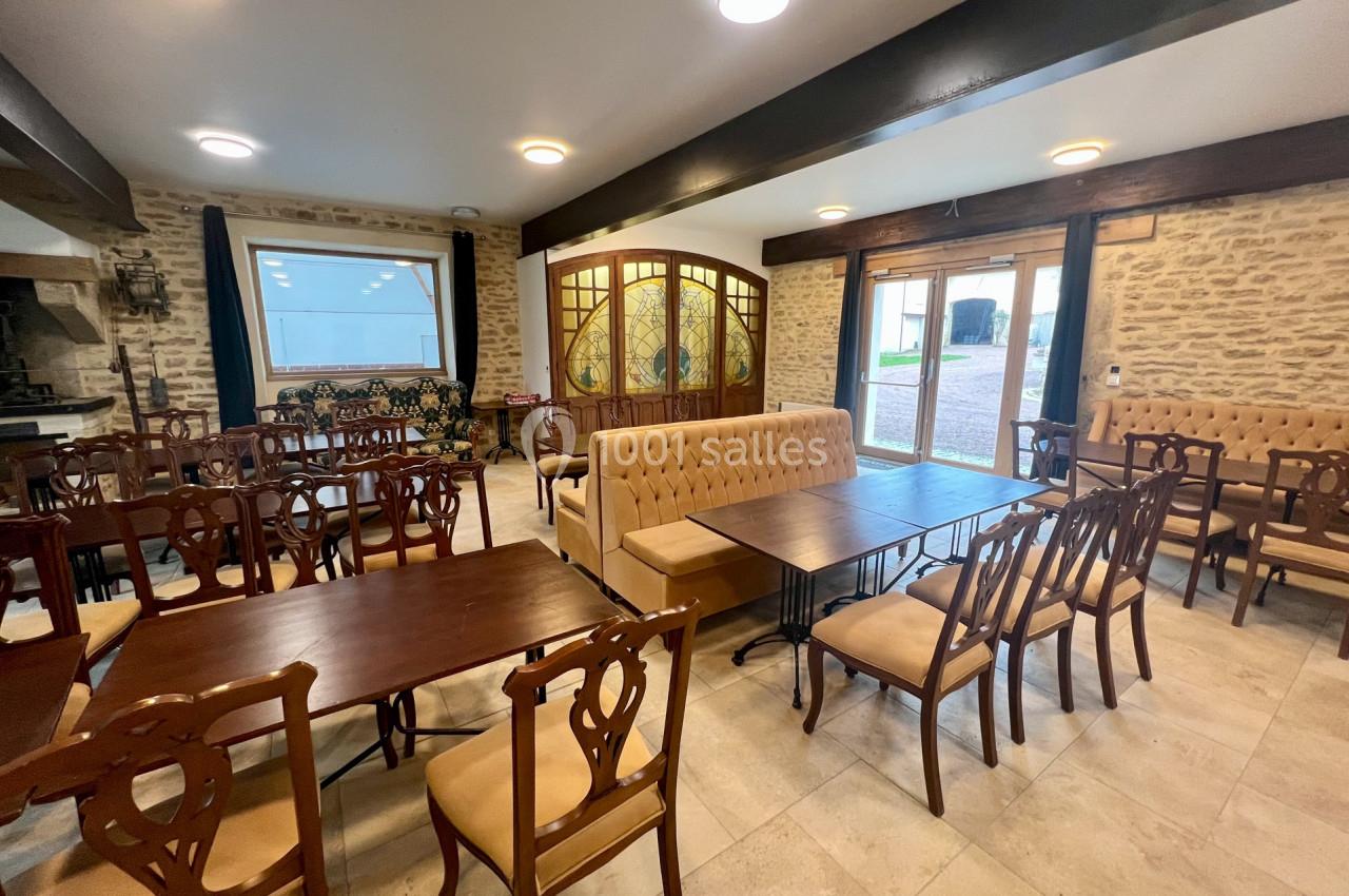 Salle lumineuse avec tables en bois, chaises rembourrées et murs en pierre, donnant sur une cour extérieure.