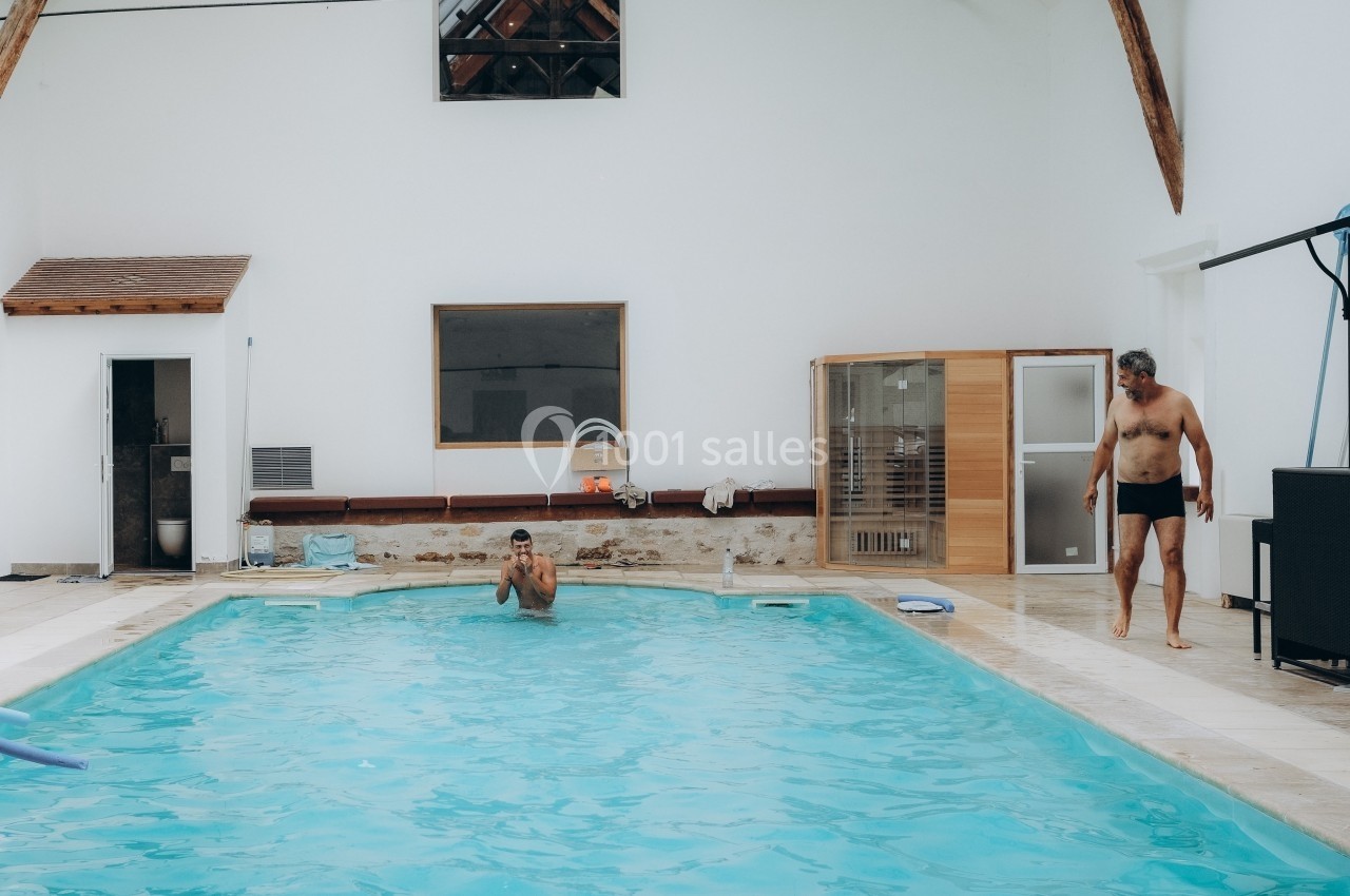 Deux hommes près d'une piscine intérieure, l'un nageant, l'autre debout sur le bord, dans un espace lumineux.